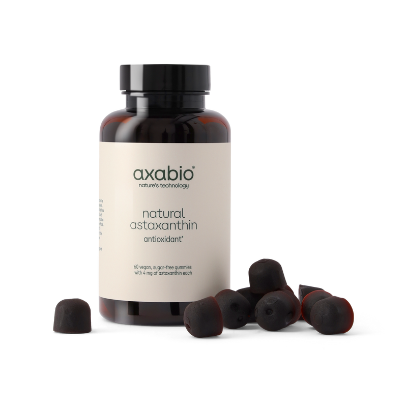 A bottle of axabio natural astaxanthin antioxidant supplement with several dark red astaxanthin capsules scattered in front, set against a solid white background.